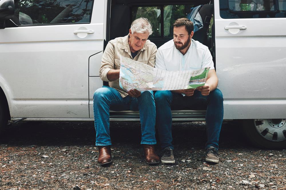 A senior man and his adult son sitting by their van, looking at a map and looking where to go