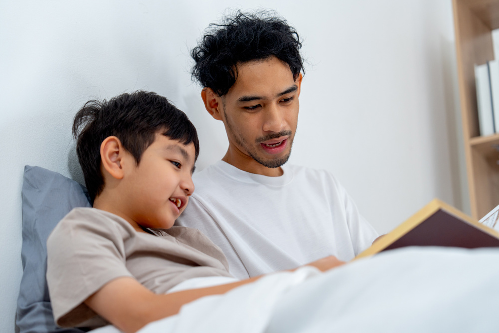 Close up view Asian young father sit near his son on bed and read the book with happiness