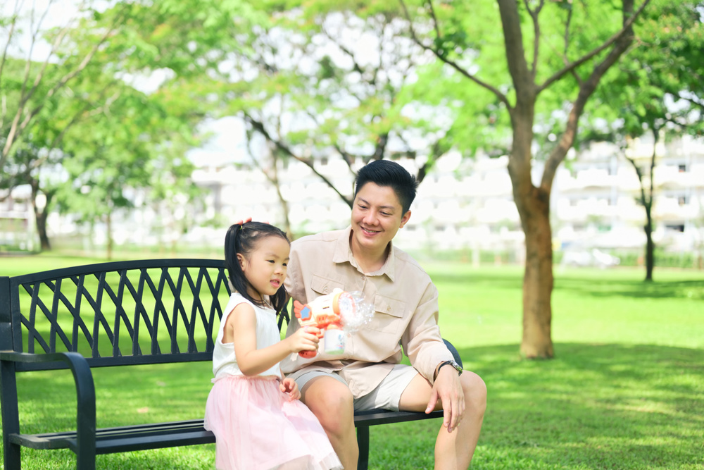 Father and daughter sit on a park bench enjoying a bubble toy on a sunny day